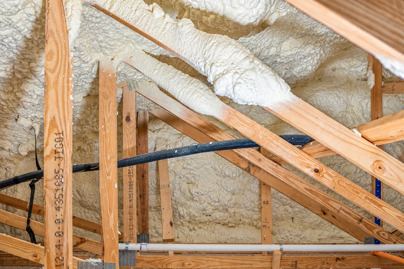 Spray foam insulation fills the roof framing and rafters around a black cable in an unfinished attic.