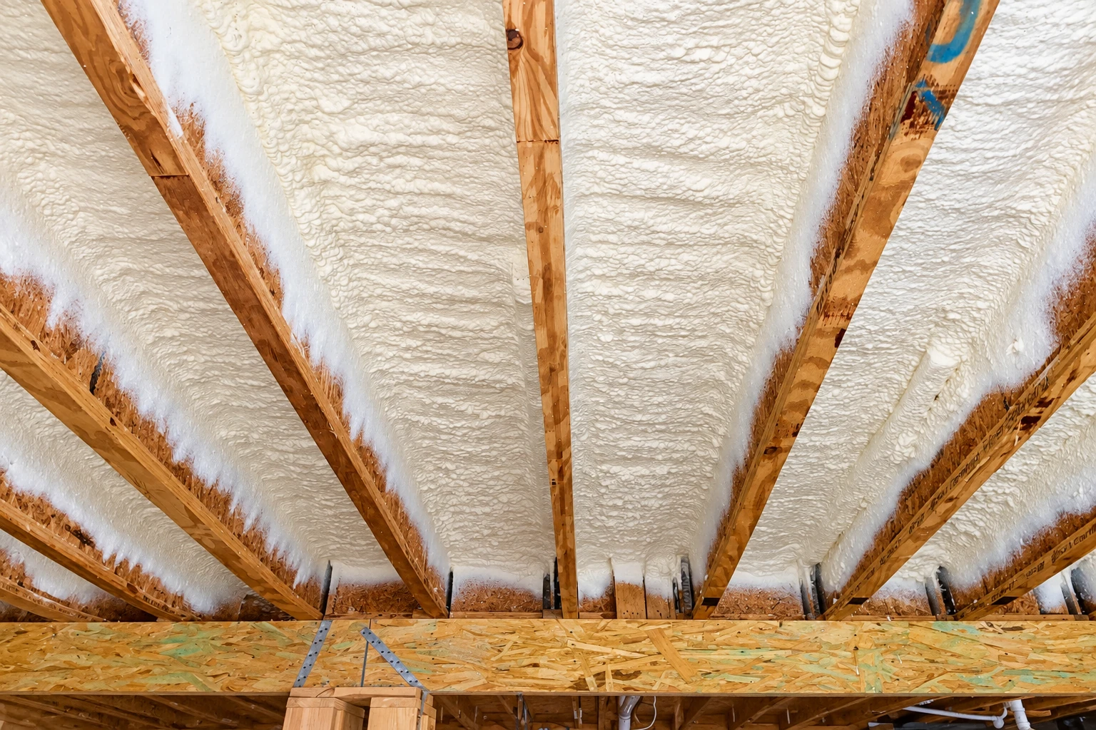 White spray foam fills the spaces between exposed wooden rafters in an attic.