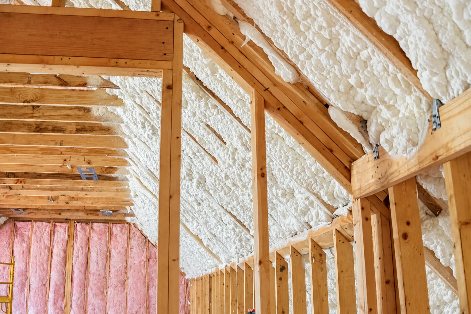 White spray foam covers the sloped roof framing in an unfinished attic, with pink batts visible along the lower wall.