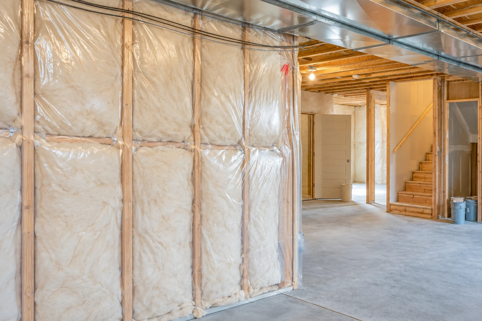 Basement wall cavities are packed with insulation behind clear plastic sheeting in an unfinished basement.