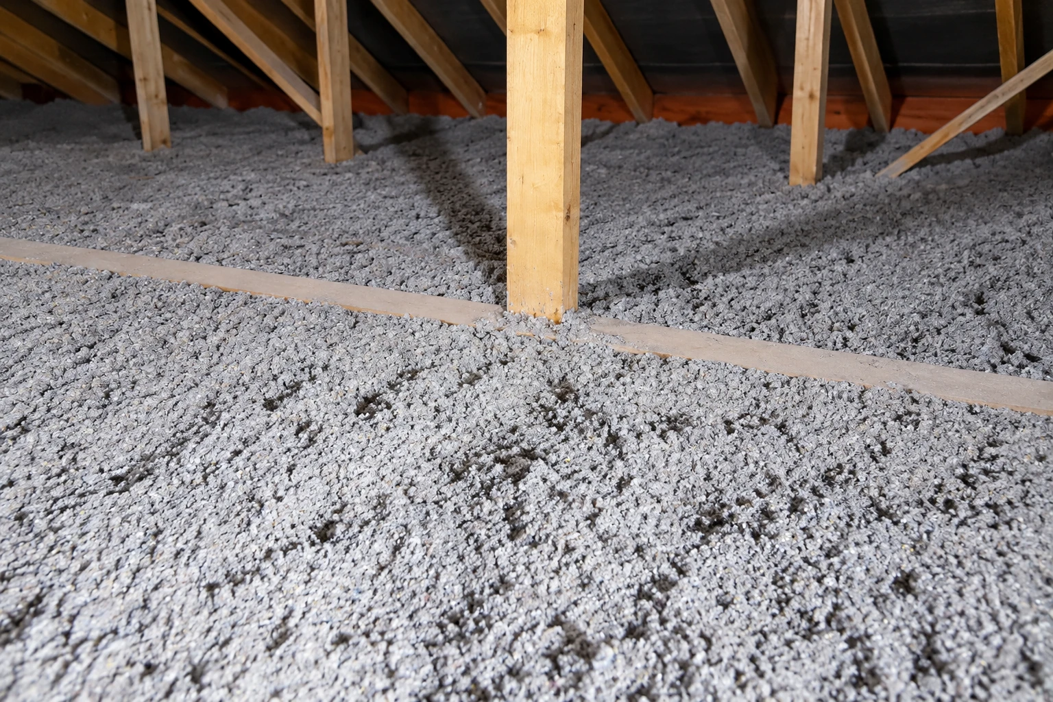 Gray blown-in insulation blankets an attic floor beneath exposed wooden roof trusses.