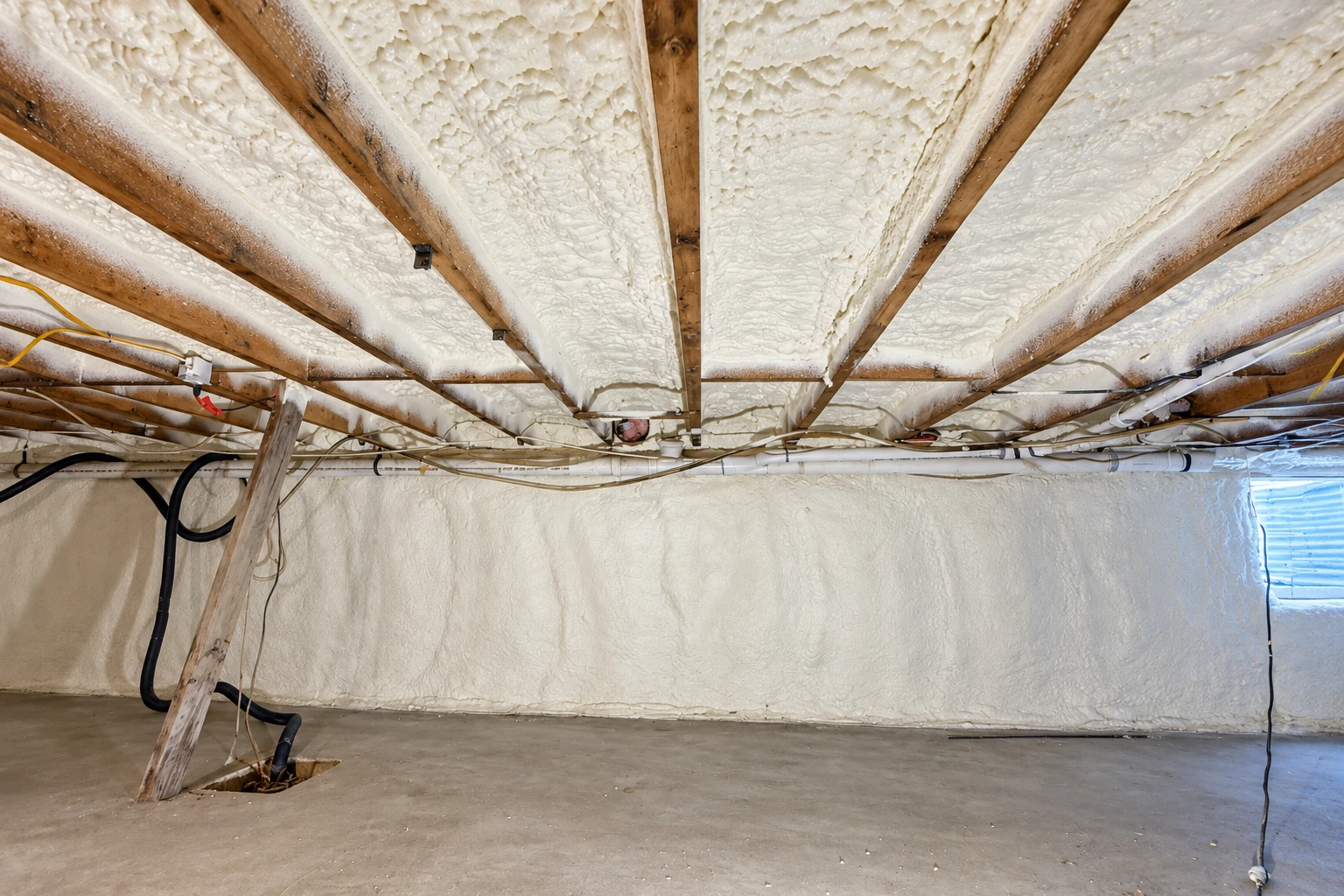 Crawl space ceiling and walls are coated with white spray foam between wooden joists, with pipes and cables running along the ceiling.