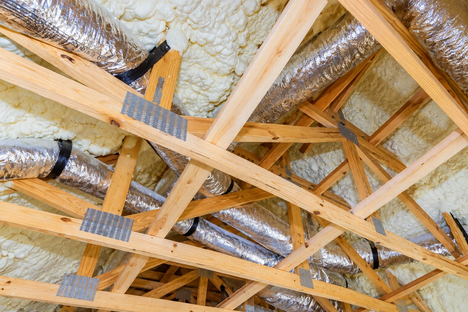 Silver flexible ducts run through a wooden attic framed and filled with white spray foam.