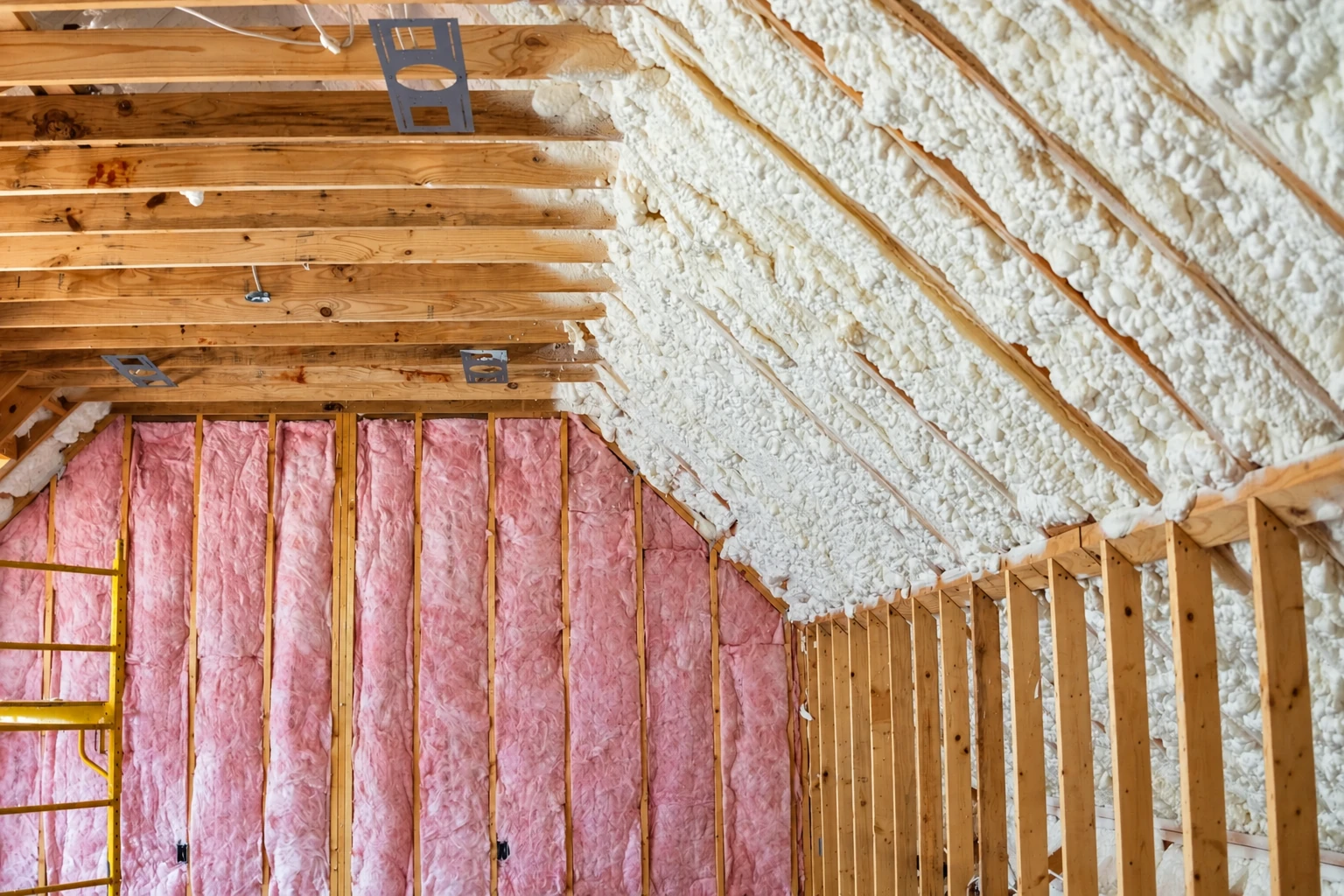 White spray foam covers a sloped attic roof beside exposed rafters and pink fiberglass insulation in the end wall.