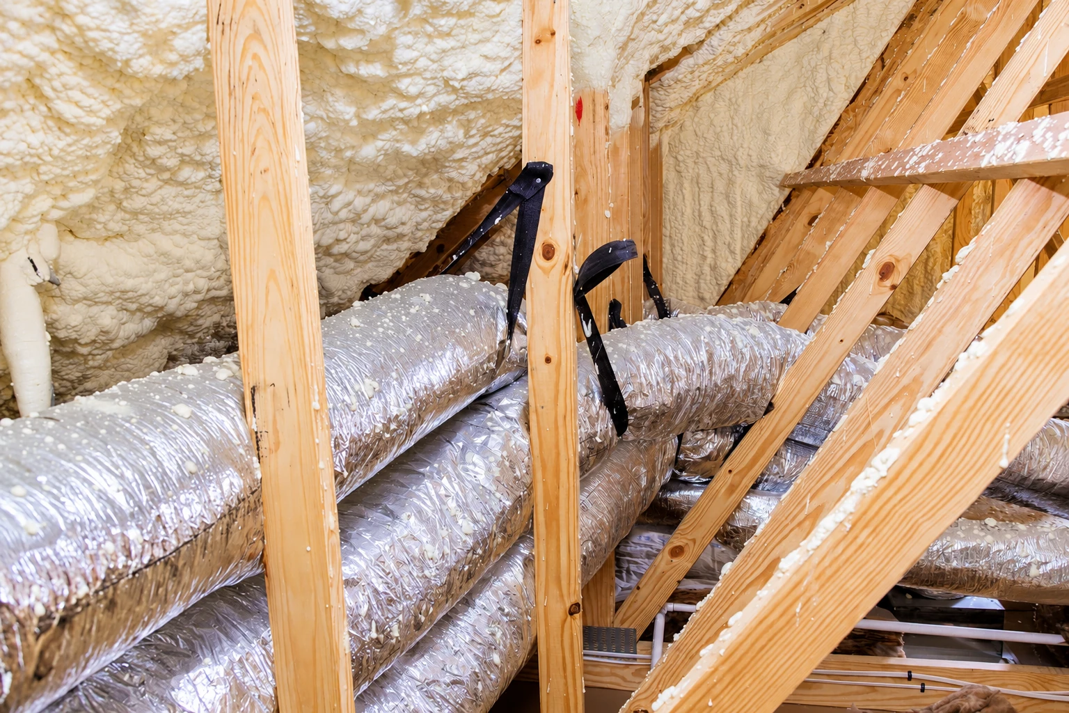 Spray foam coats the roof and walls of an attic with exposed wooden framing and foil-wrapped ducts.
